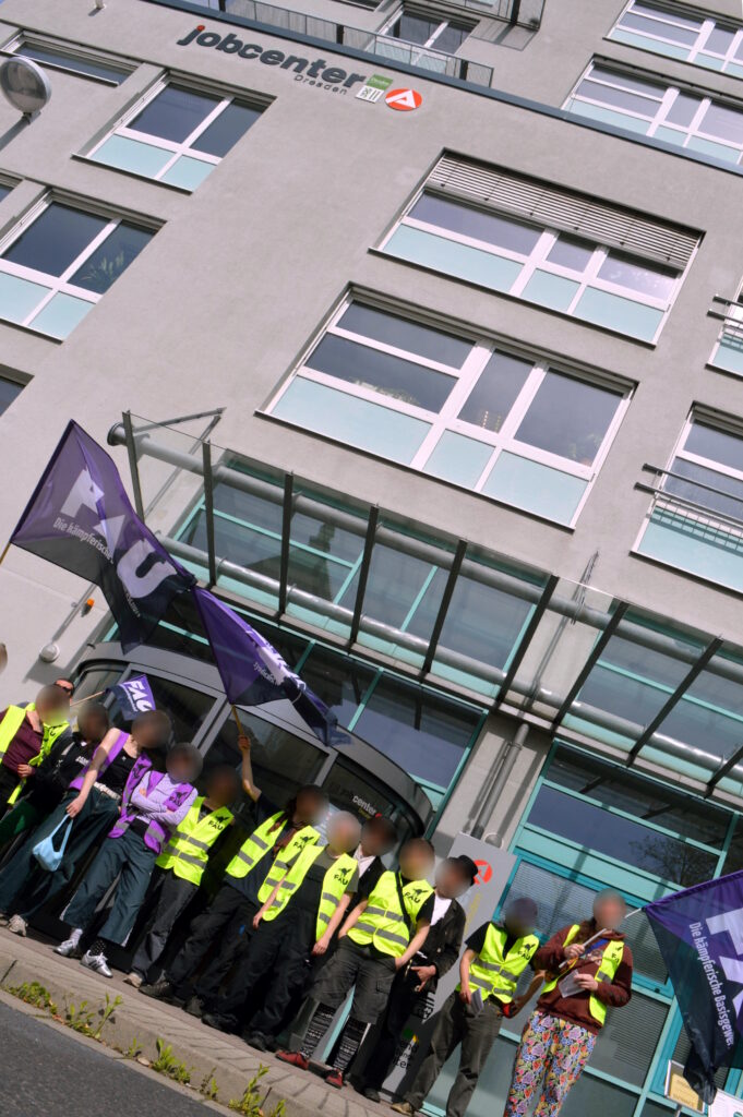 Foto mit vielen FAU-Mitgliedern vor dem Gebäude des Jobcenters. Die Personen tragen Warn-Westen und schwenken lila FAU-Fahnen.